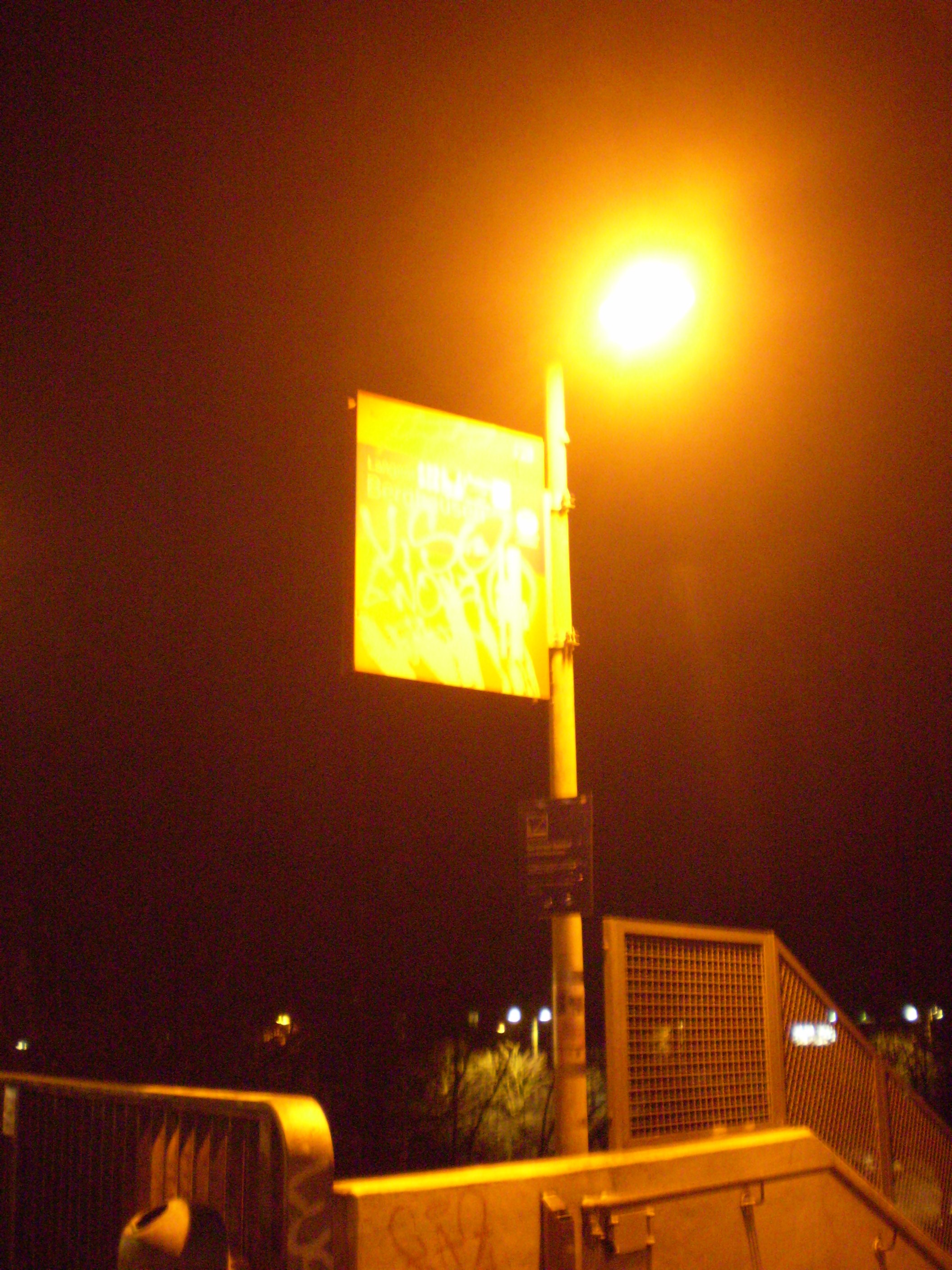 Centered is the sign of a train station. Though the lamp above it is unbearbly bright, one could confidantly assume that the author taking the picture did chose to illumintate the light to hide the location of the sign. Bright, daunting, shining light from an artifical mini sun that is made for this sign in particular. It's the begining of a staircase going down.