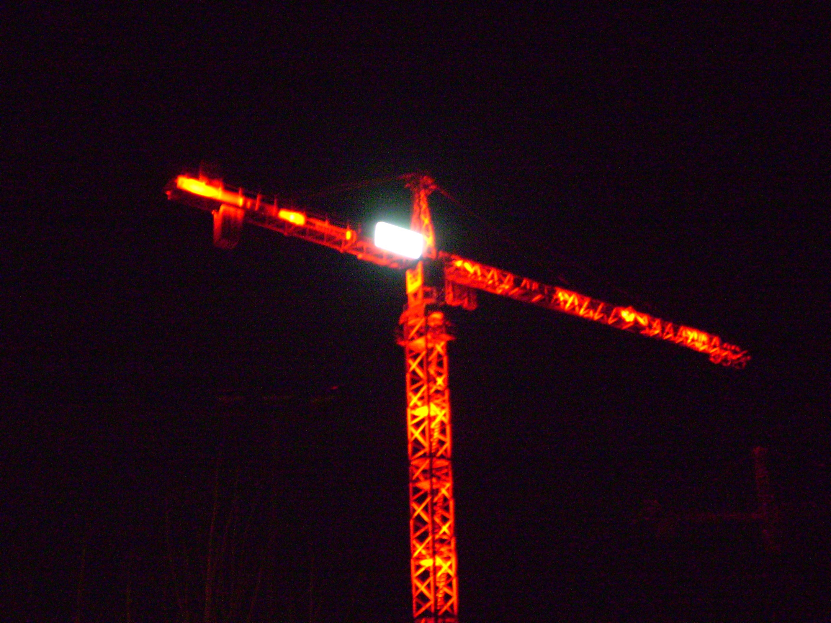A crane infront of a pitch black night. The cranes red light, that typically warns aircraft, shine in a vibrant tones tonight. Mesh with hints of yellow, the tower appears forbose and treatining, yet at the same time, like a beacon, draws one nearer.