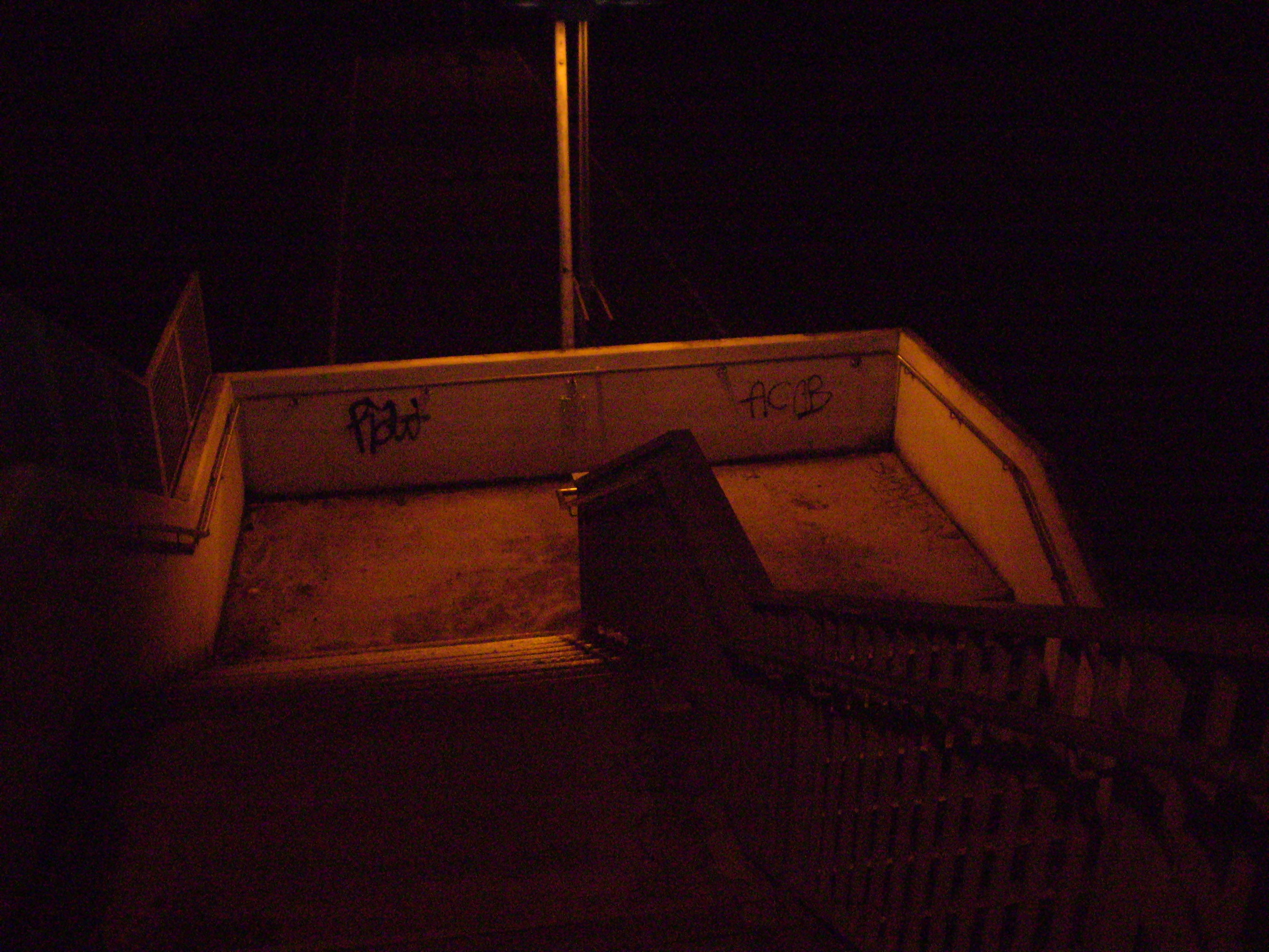 staircase with a curve going downwards. On it's walls graffity reads, 'ACAP' and with a Tag. Night shourds the dimly orange hue that envelops the walkable parts of the staircase. It's left to imagination what could be behind the stairs.